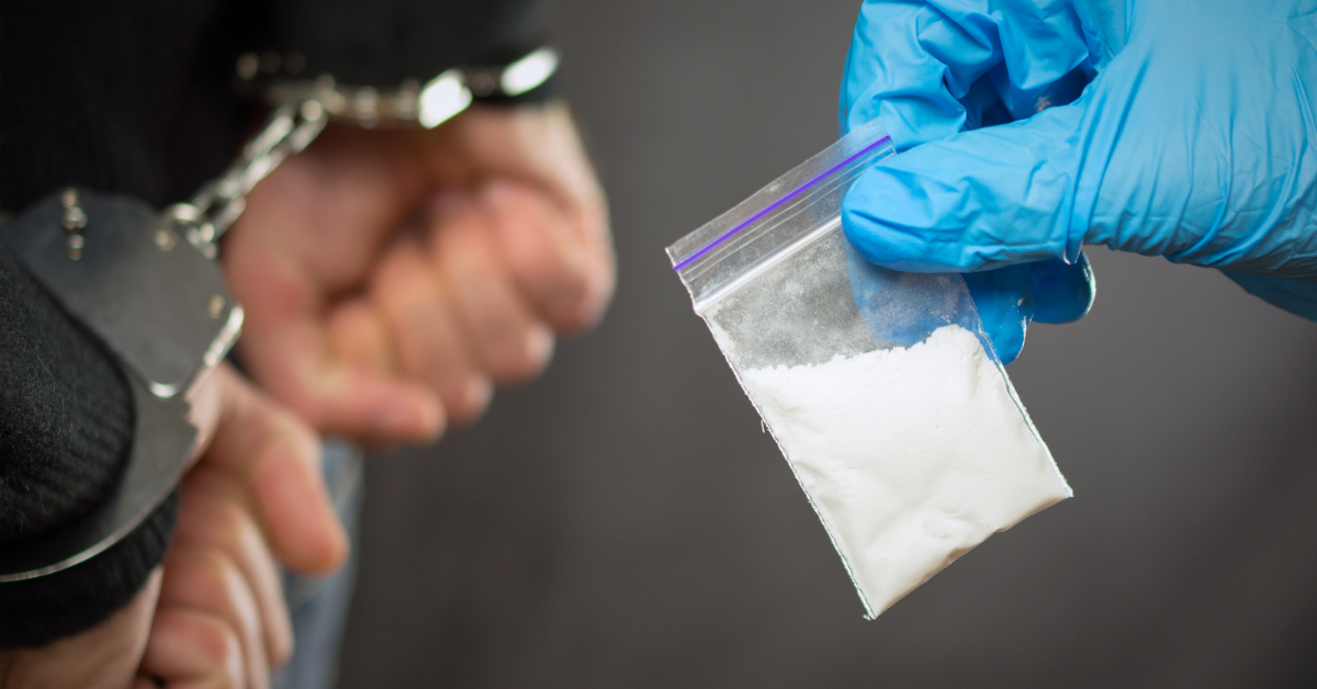 Gloved hand holding small baggie of drugs with man handcuffed in background